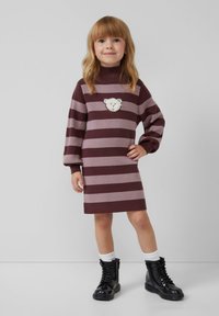Gebreide jurk met horizontale maroon en lavendel strepen, voorzien van een witte berengezichtenpatch, lange mouwen en een hoge hals. Zwarte combatboots.