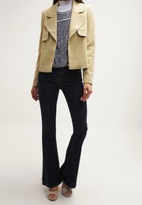 Beige fusk läderjacka med slag över en mönstrad grå topp. Mörka utställda jeans och silverfärgade sandaler med öppen tå fulländar outfiten.