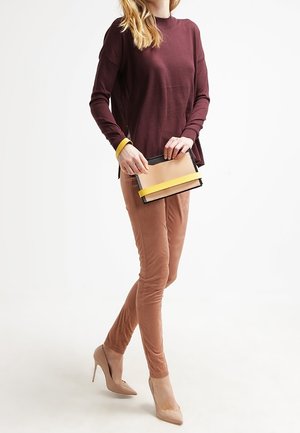 Burgundy långärmad topp, tighta beige byxor och beige klackar. Håller en flerfärgad väska med gula detaljer och svart kant.
