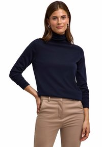 Dunkelblauer Rollkragenpullover mit langen Ärmeln und taillierter Passform, kombiniert mit beigefarbenen, figurbetonten Hosen. Weicher Stoff, minimalistischer Stil.