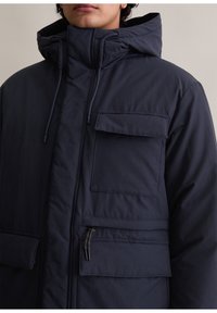 Granatowa, ocieplana kurtka z wysokim kołnierzem, regulowanym kapturem, licznymi kieszeniami i zamkiem błyskawicznym z przodu. Gładka faktura.