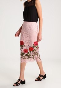 Blouse sans manches noire avec un col en dentelle texturée associée à une jupe en dentelle rose ornée de roses rouges brodées et de vignes vertes. Sandales noires.