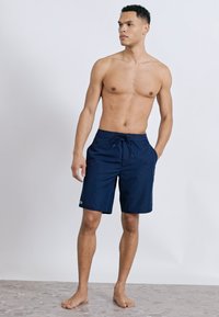 Άνδρας χωρίς πουκάμισο, όρθιος, ξυπόλητος, φορώντας σκούρα μπλε μπανάνες με ρυθμιζόμενο κορδόνι, κοιτάζει προς τα αριστερά του σε ένα απλό φωτεινό φόντο.