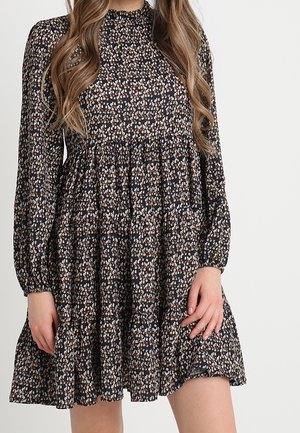 Czarna sukienka z długim rękawem, z drobnym wielokolorowym kwiatowym wzorem, wysokim kołnierzem, marszczoną talią i warstwową spódnicą, noszona przez osobę z długimi falującymi włosami.