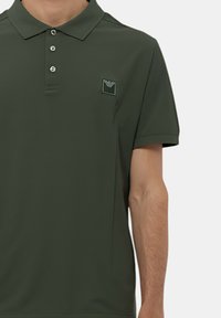 Ciemnozielona koszulka polo z kołnierzykiem, zapięciem na trzy guziki i małą naszywką z logo po lewej stronie klatki piersiowej. Materiał wydaje się gładki i lekki.