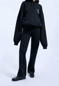 Svart oversize-hoodie med framficka, kombinerad med svarta utsvängda byxor och svarta loafers. Materialet verkar mjukt med en slät textur.