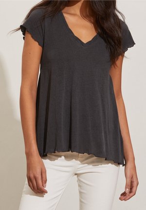 Kobieta w luźnej, ciemnoszarej koszulce z krótkim rękawem i dekoltem w serek z falbankami, w połączeniu z białymi spodniami na jednolitym tle.