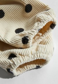 Romig geribbelde stof met zwarte stippen, met elastische randen. De textuur is zacht en het ontwerp is casual en lichtgewicht.