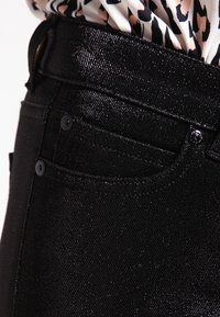 Svarta denimjeans med glänsande textur och två framfickor. Har knäppning med knappar och hög midja. Nära vy av tygdetaljer.