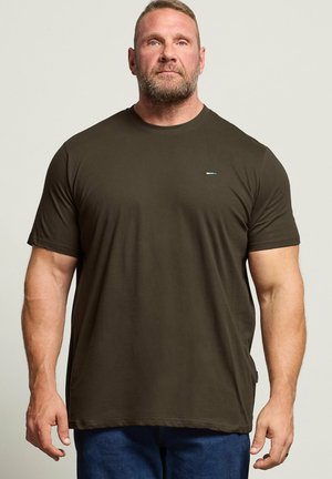 Muskeløs mand med kort hår og skæg iført mørke olivengrøn T-shirt og blå jeans, stående mod en ensfarvet lys baggrund.