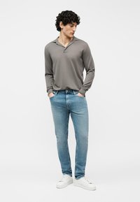 Νέος άνδρας με σγουρά μαλλιά, που φοράει γκρι πολύ μακρυμάνικη μπλούζα πόλο, ανοιχτό μπλε στενά τζιν και άσπρα αθλητικά παπούτσια, στέκεται με τα χέρια στις τσέπες.