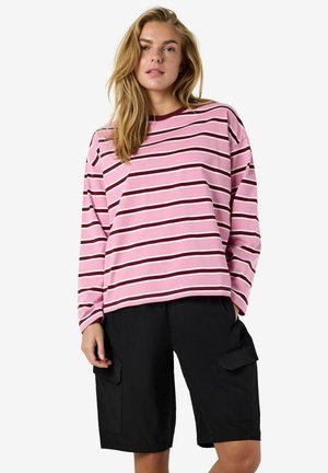 Femme aux cheveux longs et blonds portant un haut à manches longues rayé rose, blanc et bordeaux, ainsi qu'un short cargo noir jusqu'aux genoux, debout devant un fond blanc.