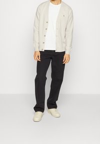 Cárdigan beige con botones, usado sobre una camiseta blanca, combinado con pantalones negros y zapatillas de color claro. Diseño sencillo y textura suave.
