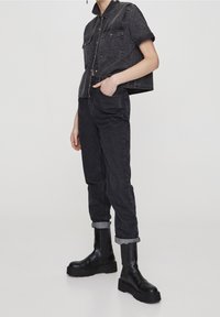 Chemise courte en denim noir à manches courtes, avec deux poches avant. Pantalon en denim foncé fuselé avec revers. Porté avec des bottines noires épaisses.