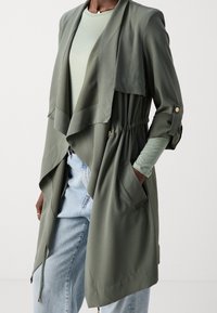 Langer, olivgrüner, leichter Mantel mit drapiertem Vorderteil, Kordelzug in der Taille und zwei Seitentaschen; kombiniert mit einem hellgrünen Oberteil und Jeans.