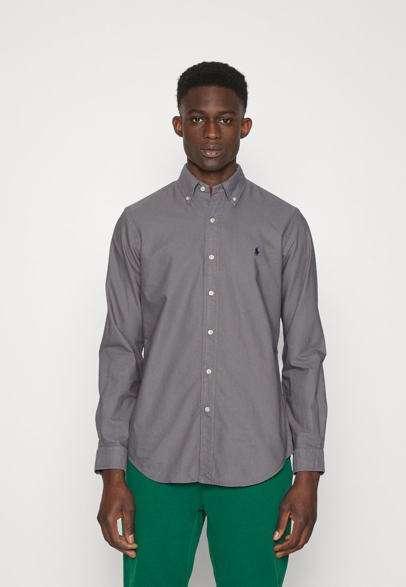 Camisa de botones de manga larga gris con cuello, que presenta un pequeño logo azul en el pecho. Combinada con pantalones verdes. Textura suave.