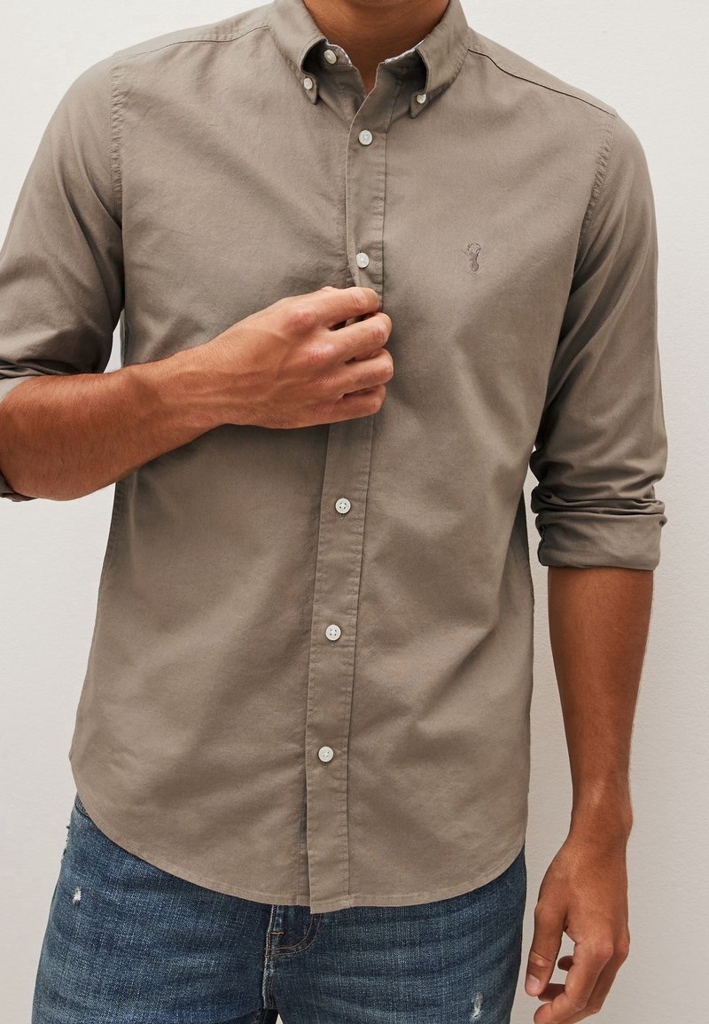 Homme boutonnant une chemise marron clair à manches longues retroussées, portant un jean bleu légèrement usé.