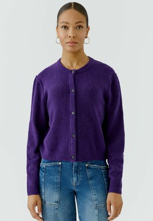 Frau mit zurückgekämmtem Haar, die goldene Creolen, eine lila geknöpfte Strickjacke und blaue Jeans mit großen Fronttaschen trägt.