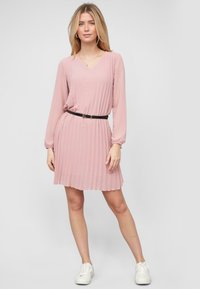 Abito rosa a pieghe con maniche lunghe, scollatura a V e vita cintrata accentuata da una cintura nera. Indossato con sneakers bianche. Texture liscia.