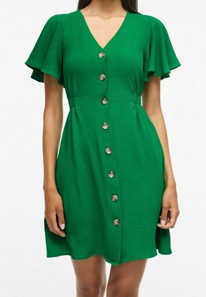 Femme portant une robe courte verte avec des manches flottantes, des boutons sur le devant et une taille légèrement froncée.