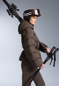 Hnedá puffa bunda so priliehavým pásom, kapucňou a prešívanou textúrou; nosí hnedú lyžiarsku prilbu s okuliarmi; drží čierne lyžiarske palice.