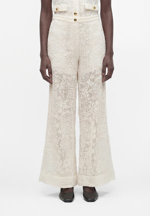Pantalon large en dentelle crème avec broderies florales porté avec une veste courte assortie et des chaussures noires à bout carré.
