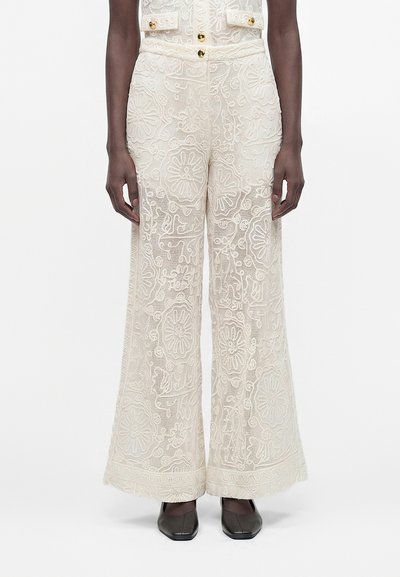 Pantalon large en dentelle crème avec broderies florales porté avec une veste courte assortie et des chaussures noires à bout carré.