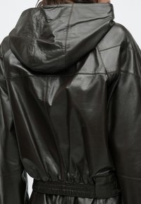 Detailný záber na osobu nosiacu tmavohnedú lesklú koženú bundu s veľkou kapucňou a elastickým pásom.