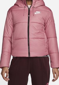 Rosa puff-jacka med huva, quiltad design och svart dragkedja. Har en vit Nike-logotyp på bröstet och elastiska ärmslut.