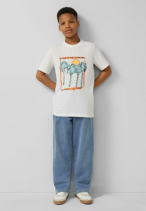 Boy standing against gray wall wearing white t-shirt with palm tree and sun print, light blue wide-leg jeans, and white sneakers.