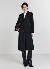 Schwarzer langer Mantel aus strukturiertem Stoff mit Reverskragen, vorderen Knöpfen und zwei Pattentaschen. Kombiniert mit glänzenden schwarzen Lederhosen.