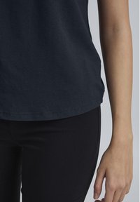 Dunkelblaues Baumwoll-T-Shirt mit lässiger Passform. Der Saum ist leicht kurvenförmig. Getragen mit schwarzen enganliegenden Hosen. Glatte Textur, minimalistische Details.