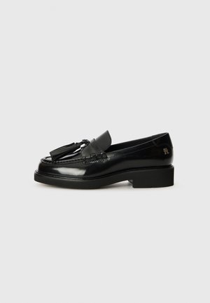 Mocassins em couro preto com um acabamento brilhante, apresentando um detalhe de franja, um acabamento costurado e um tacão baixo. Logótipo em relevo no tacão.