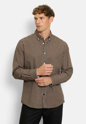 Man adjusting cuff on brown, black, and white checkered button-up shirt, standing against a plain light background.