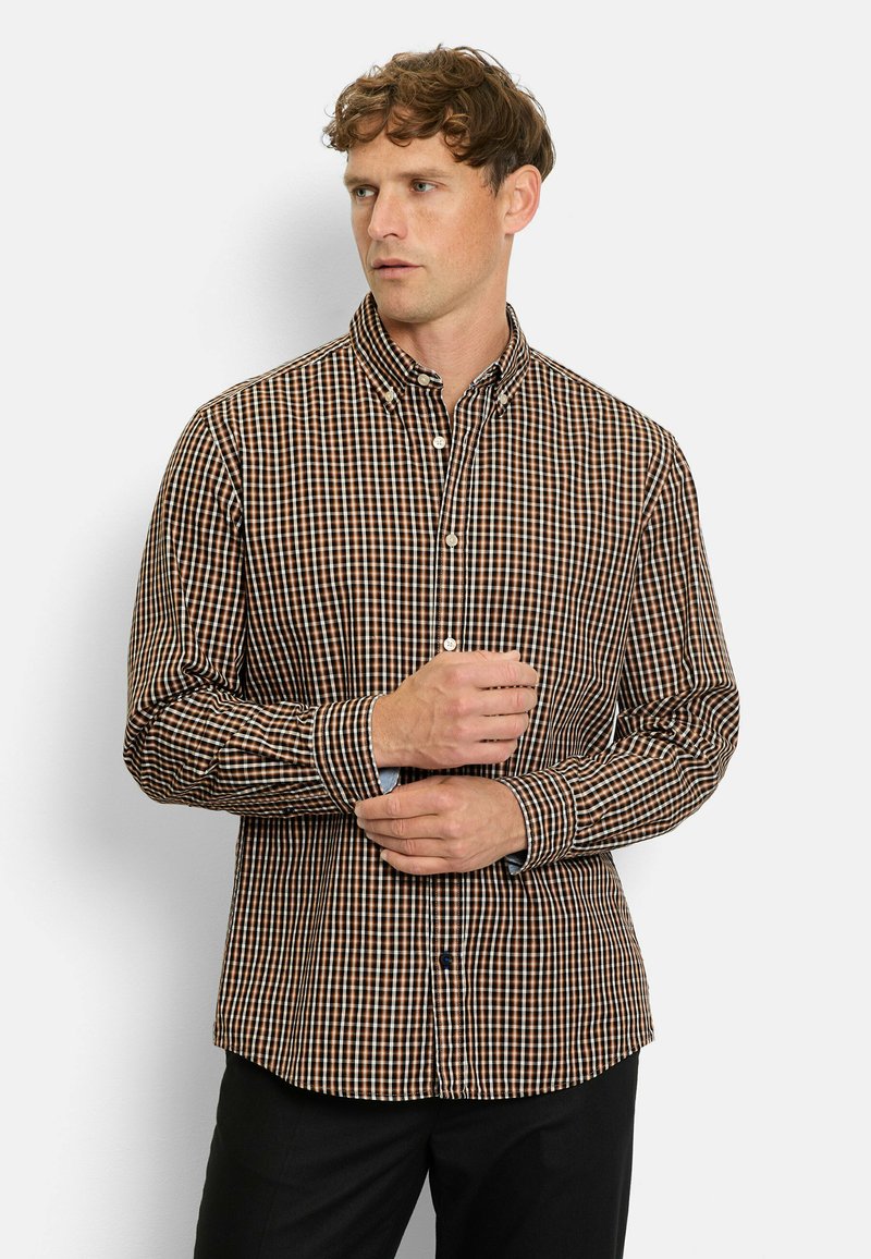 Mann, der das Bündchen eines braun-, schwarz- und weißkarierte Hemds mit Knöpfen anpasst, steht vor einem einfarbigen hellen Hintergrund.