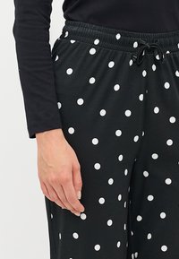 Pantaloni negri cu buline, cu talie elastică, detaliu cu șnur și modele circulare albe distribuite uniform pe întreaga țesătură.