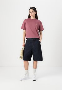 Rosa t-shirt med kort ärm, mörka jeansshorts, vita högskurna sneakers, vita strumpor. Håller en beige väska. Enkel design och avslappnad passform.