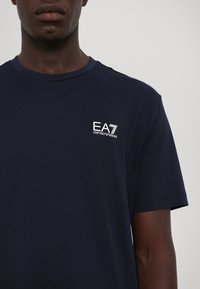 Tamsiai mėlynas medvilninis marškinėliai su baltu EA7 Emporio Armani logotipu ant krūtinės, apvaliu kaklu ir trumpomis rankovėmis, su glotnia tekstūra.