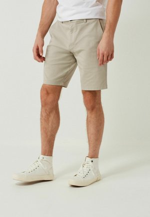 Mand iført beige shorts til knæene, hvide high-top sneakers og en hvid skjorte, stående mod en ensfarvet lys baggrund.