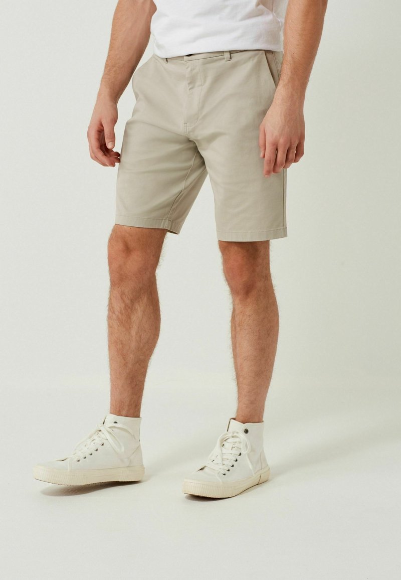Homme portant un short beige jusqu'aux genoux, des baskets montantes blanches et une chemise blanche, debout devant un fond clair uni.