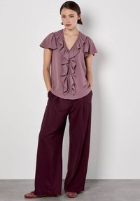 Lila geraffte Bluse mit V-Ausschnitt und kurzen Ärmeln, kombiniert mit lockeren burgunderfarbigen Hosen und hellrosa Ballerinas, was einen entspannten Look erzeugt.