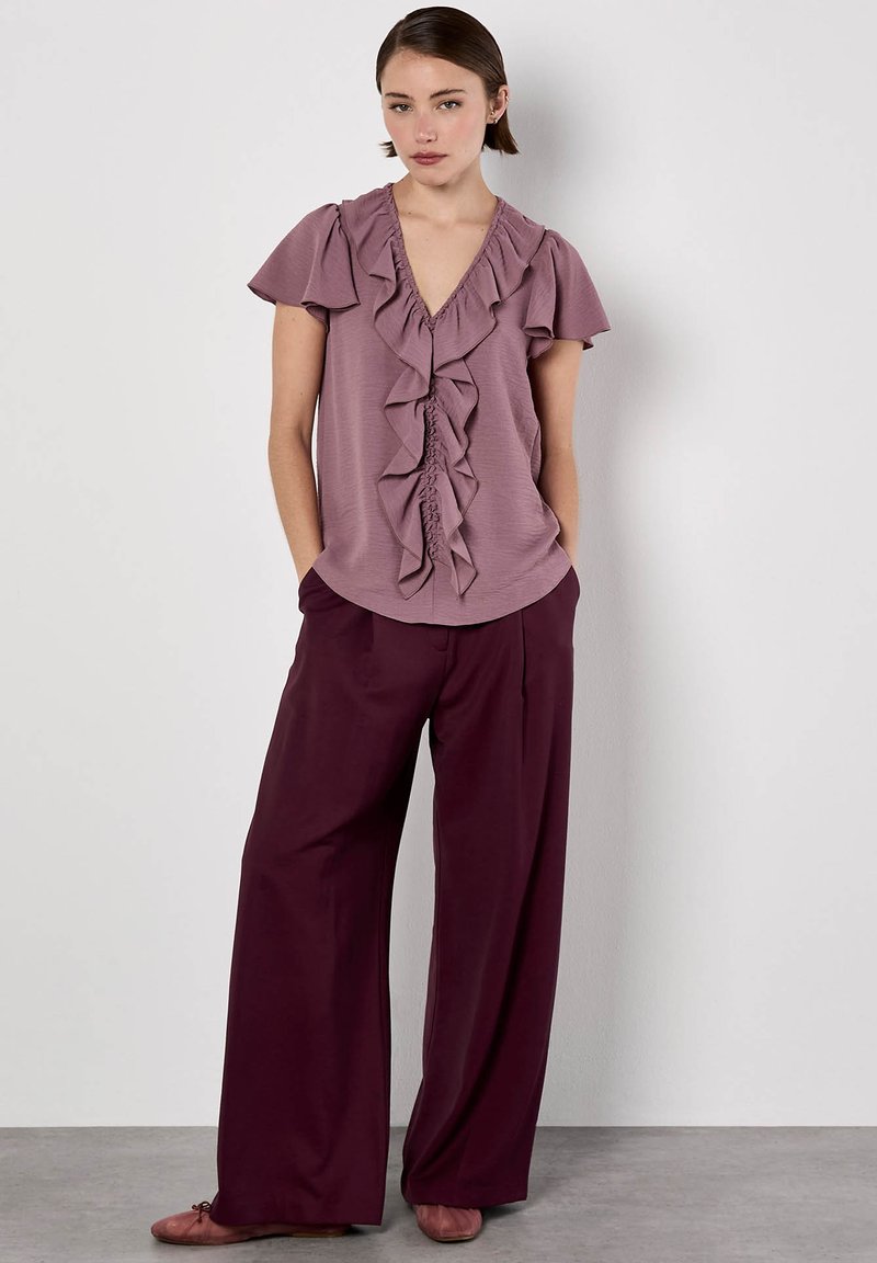 Lila geraffte Bluse mit V-Ausschnitt und kurzen Ärmeln, kombiniert mit lockeren burgunderfarbigen Hosen und hellrosa Ballerinas, was einen entspannten Look erzeugt.
