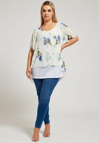 Blusa floreale semi-trasparente bianca con motivo a tulipani blu, abbinata a un top a strati grigio e jeans skinny blu scuro. Sandali con tacco trasparente.