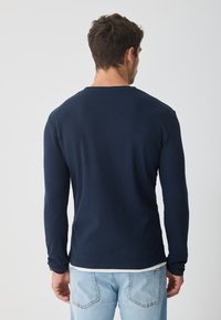 Temno modra majica z dolgimi rokavi in rebrasto teksturo, kroja s čolnasto ovratnikom, v kombinaciji s svetlo modrimi džinsami, prikazana s hrbtne strani.