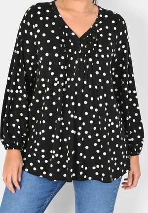 Blusa nera con pois bianchi, scollo a V, parte frontale plissettata e maniche a sbuffo. Realizzata in tessuto morbido, abbinata a jeans blu.
