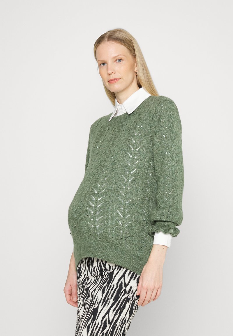Pulôver verde tricotado com padrão em chevron, gola redonda e mangas balão, combinado com calças listradas em zebra preto e branco.