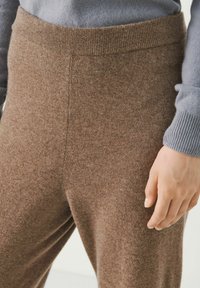 Pantalones de punto marrón con una textura suave y una cinturilla elástica ancha. Un suéter de color azul claro parcialmente visible en la parte superior del brazo.
