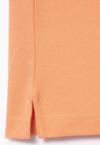 Camisa polo de manga corta, fabricada en tejido texturizado de color coral claro, presenta aberturas en los laterales del dobladillo, con un detalle de costura visible.