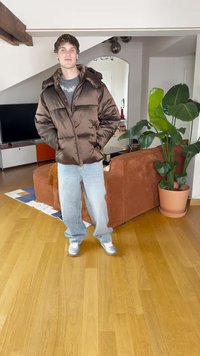 Καφέ μπουφάν με κουκούλα, ανοιχτό μπλε τζην και γκρι sneakers. Εσωτερικός χώρος με ξύλινο δάπεδο και πράσινο φυτό σε γλάστρα.