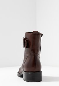 Bottine en cuir marron avec bout rond, détails cousus, fermeture éclair latérale et sangle décorative à l'arrière. Talon carré bas.